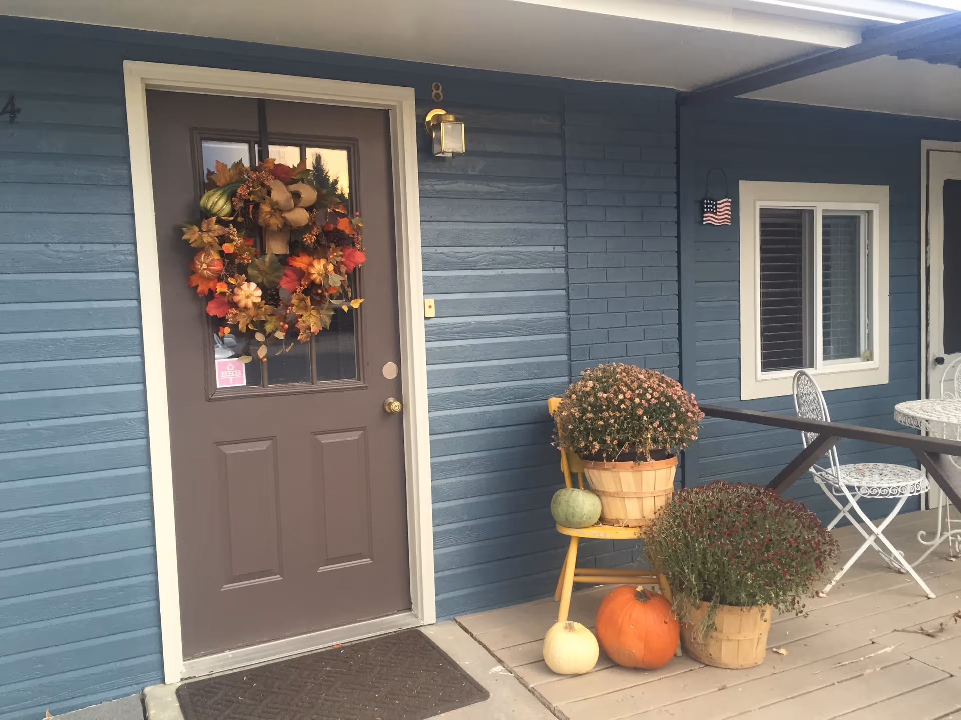 Blue exterior front door with an autumn wreath, pumpkins and potted mums on the porch beside a small table and chairs.