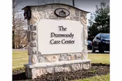 Stone monument sign that reads "The Dunwoody Care Center" on a grassy area with parked cars in the background.