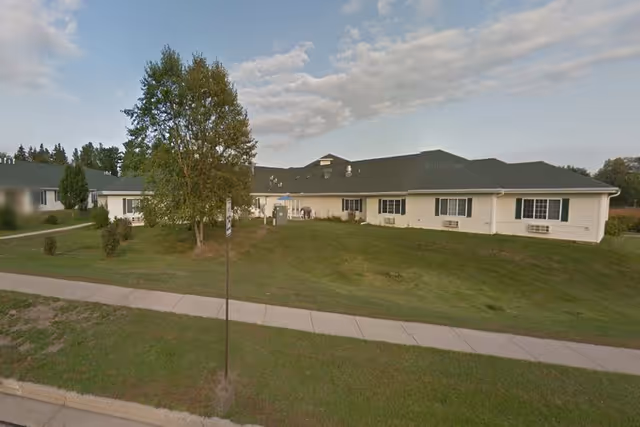 Exterior view of a single-story senior living facility building with a green roof and beige siding, surrounded by grass and a few trees under a partly cloudy sky.