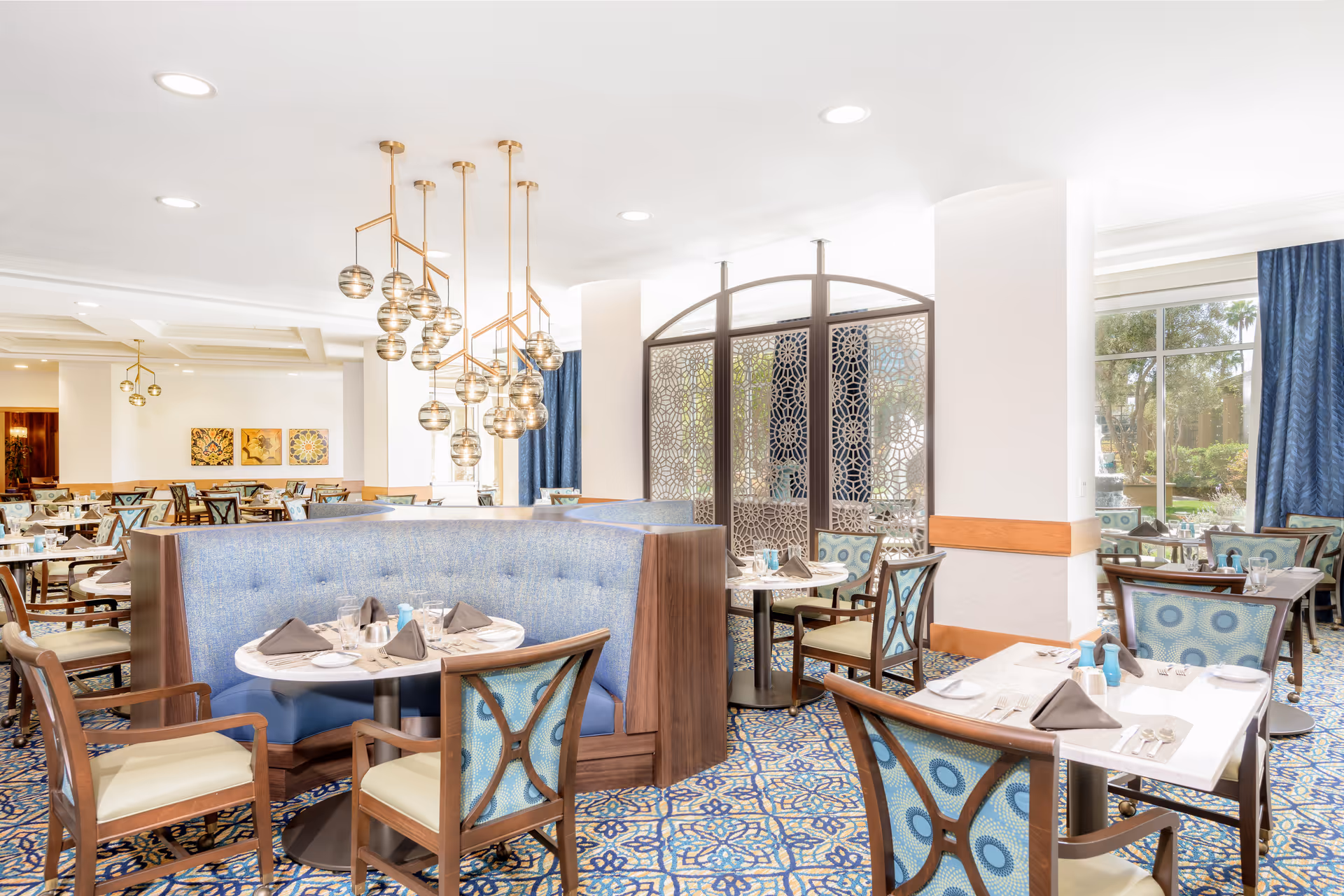 Bright and elegant dining room with blue patterned carpet and chairs, round and square tables set with plates, glasses, and napkins. A curved blue booth seating area is centrally located under modern pendant lights. Large windows with blue curtains allow natural light and a view of greenery outside. Decorative metal screen panels separate parts of the room.