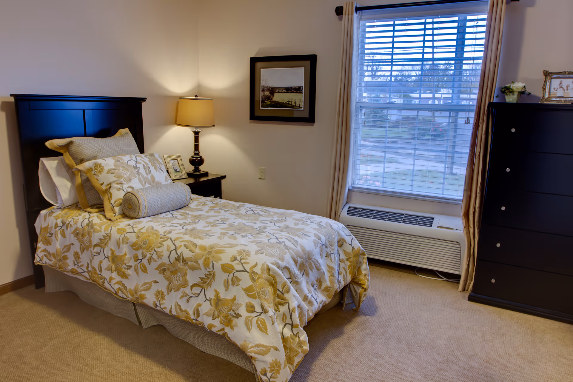 A neatly made single bed with floral patterned bedding in shades of yellow and beige, a bedside table with a lamp and a framed photo, a window with blinds and curtains, and a black dresser with a small plant and framed picture on top in a cozy bedroom.