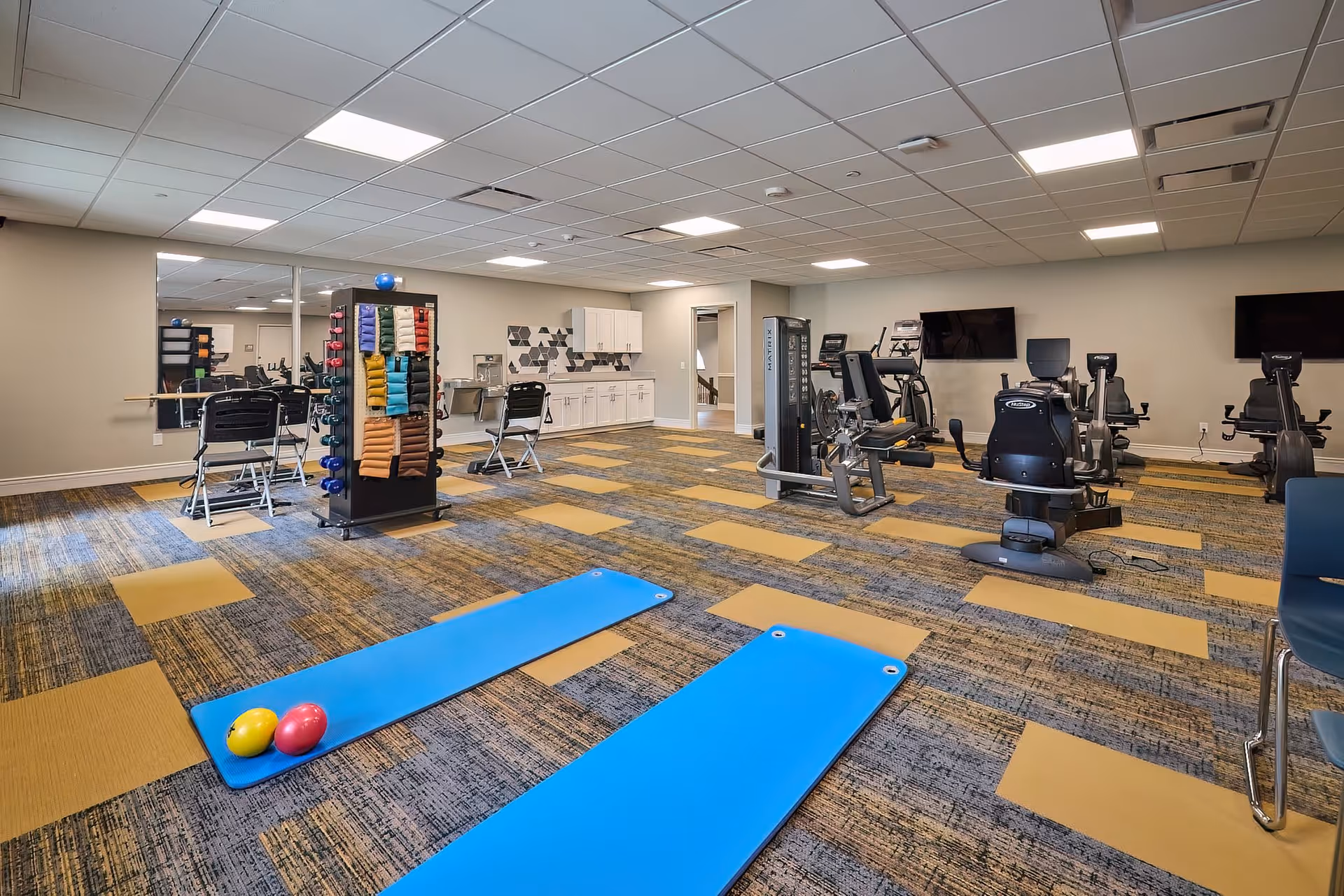 A spacious fitness room with exercise equipment including stationary bikes, weight machines, and chairs. Two blue exercise mats with small colorful balls are placed on the patterned carpet floor. The room has a drop ceiling with fluorescent lighting, a wall-mounted TV, a rack with various colored towels, and a mirrored wall on one side.