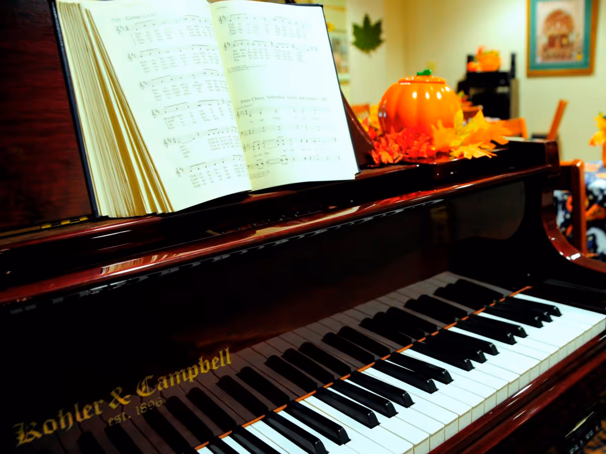 Close-up view of a polished Kohler & Campbell piano keyboard with an open sheet music book on the music stand. The piano is decorated with autumn-themed items including a small pumpkin and fall leaves. In the background, there are chairs and framed pictures on the wall, suggesting a cozy indoor setting.