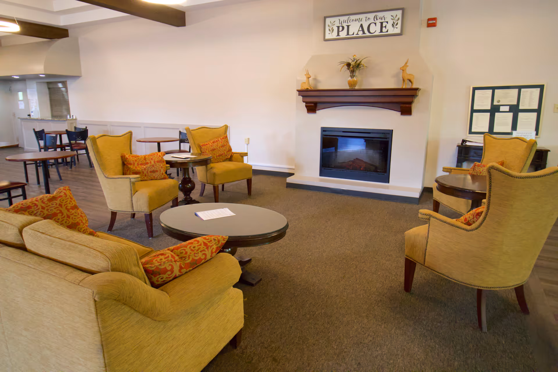 A cozy living room area with beige upholstered chairs and a sofa adorned with patterned orange cushions. There are several round wooden tables placed between the seating. A fireplace is set into the wall with a wooden mantel above it, decorated with small sculptures and a vase with flowers. Above the mantel is a sign that reads 'Welcome to Our Place'. In the background, there are additional tables and chairs, and a bulletin board on the wall.