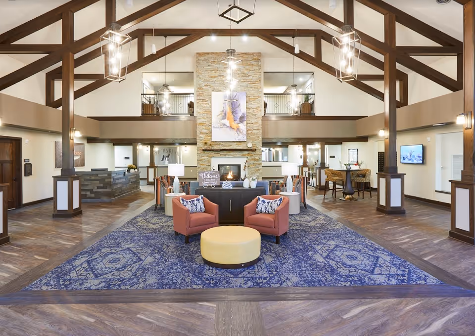 Spacious senior living lobby with vaulted wooden beams, seating arranged around a central fireplace and a large blue area rug.
