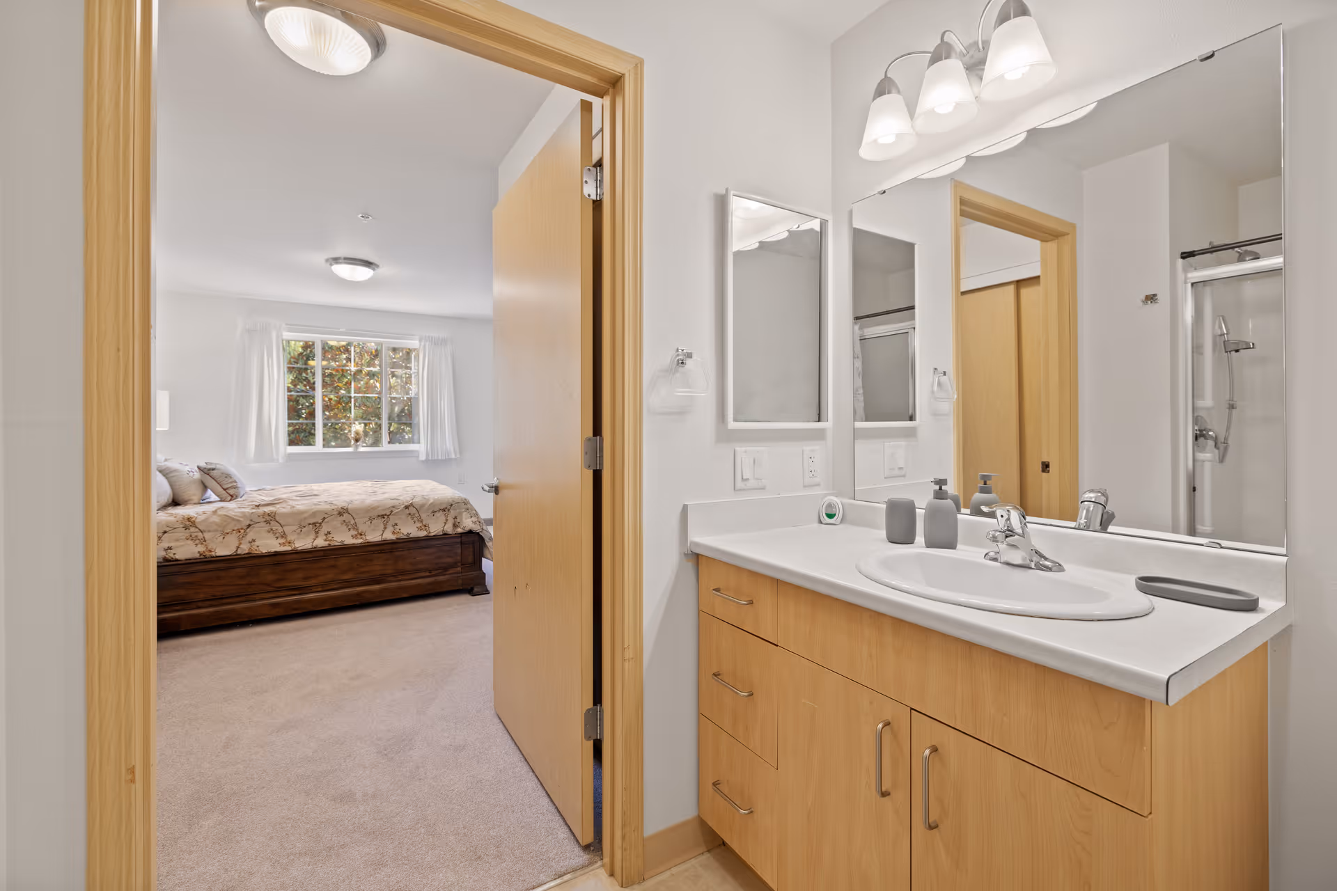 View of a bathroom vanity with a sink, soap dispensers, and a large mirror. The bathroom door is open, showing a bedroom with a bed, pillows, a window with white curtains, and ceiling lights.