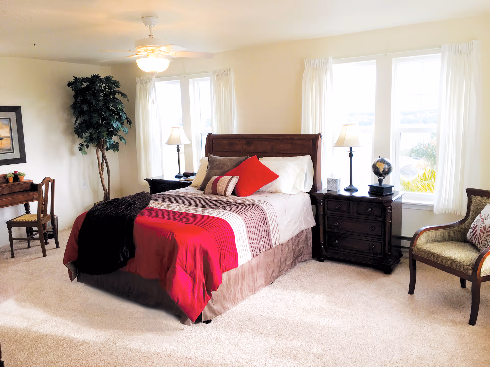 A bright and cozy bedroom with a large bed covered in a red and beige striped comforter and multiple pillows. There are two dark wooden nightstands on either side of the bed, each with a lamp. A globe and a small decorative box are on the right nightstand. The room has three large windows with white curtains, a ceiling fan with a light, a wooden chair with a cushion, a small desk with a chair, and a tall potted plant in the corner.