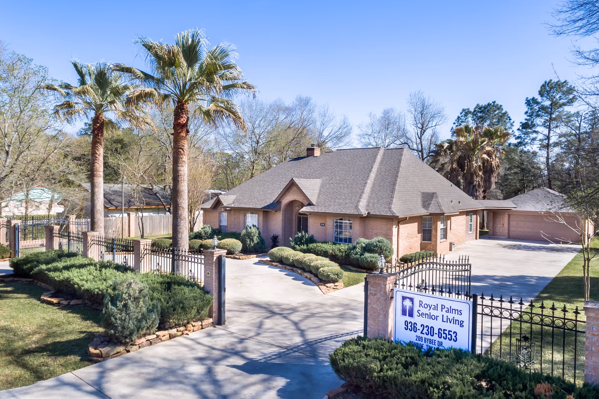 Exterior view of a single-story brick building with a gated driveway and well-maintained landscaping including palm trees and shrubs under a clear blue sky. A sign near the gate reads 'Royal Palms Senior Living' with contact information.