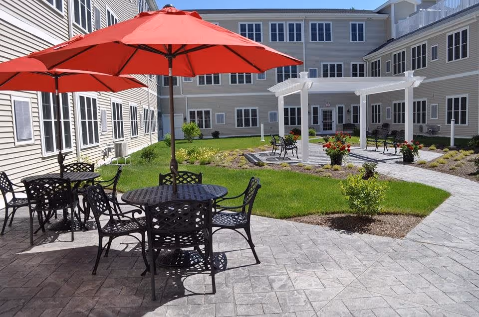 Outdoor courtyard area at an assisted living facility with round metal tables and chairs under red umbrellas, a paved walkway, green grass, flower beds, and a white pergola with benches.