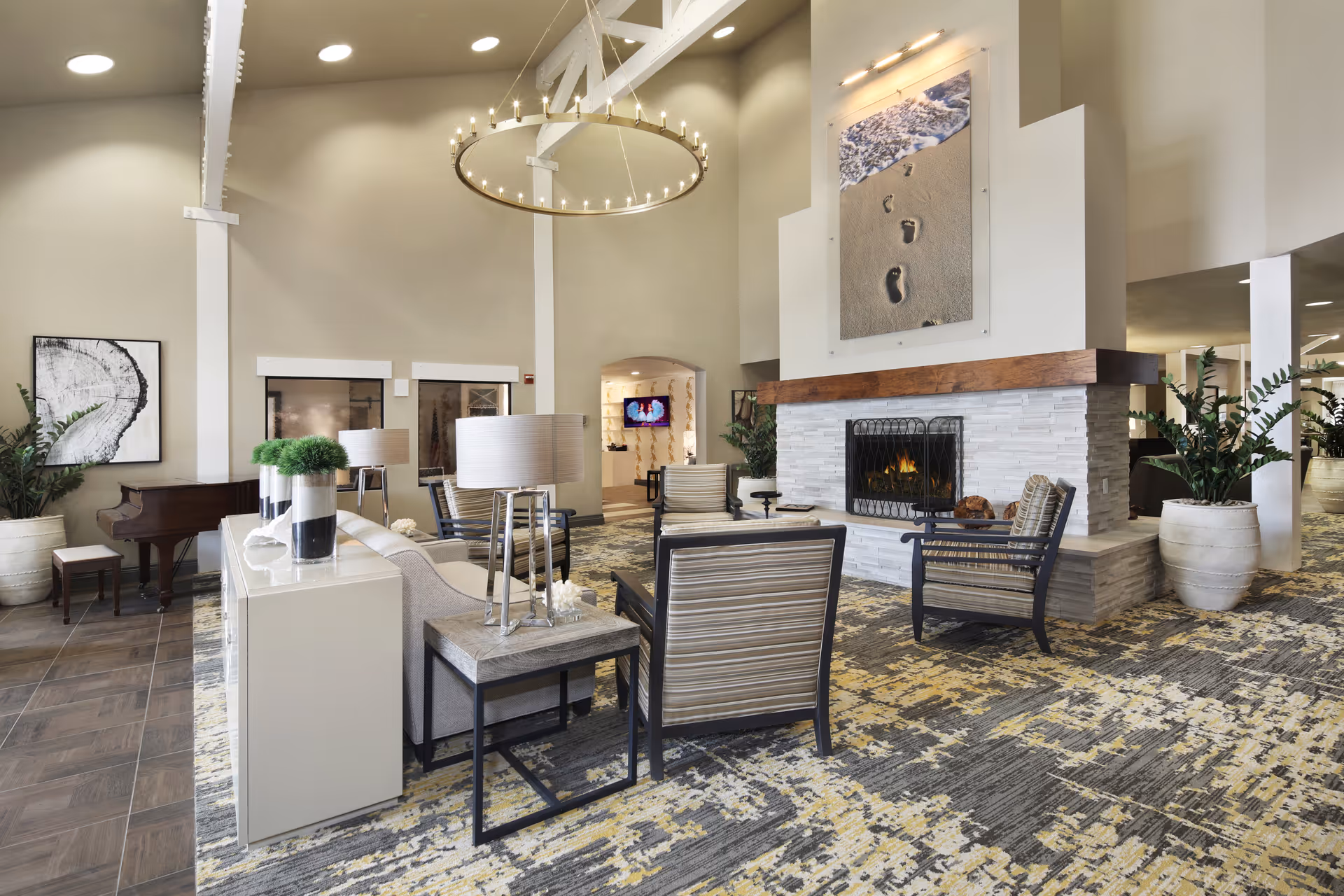 A spacious and well-lit living room area in a senior living facility featuring a modern fireplace with a large artwork of footprints in the sand above it. The room has several comfortable chairs and sofas arranged around the fireplace, a grand piano in the corner, potted plants, and a large circular chandelier hanging from the ceiling. The floor is covered with a patterned carpet.
