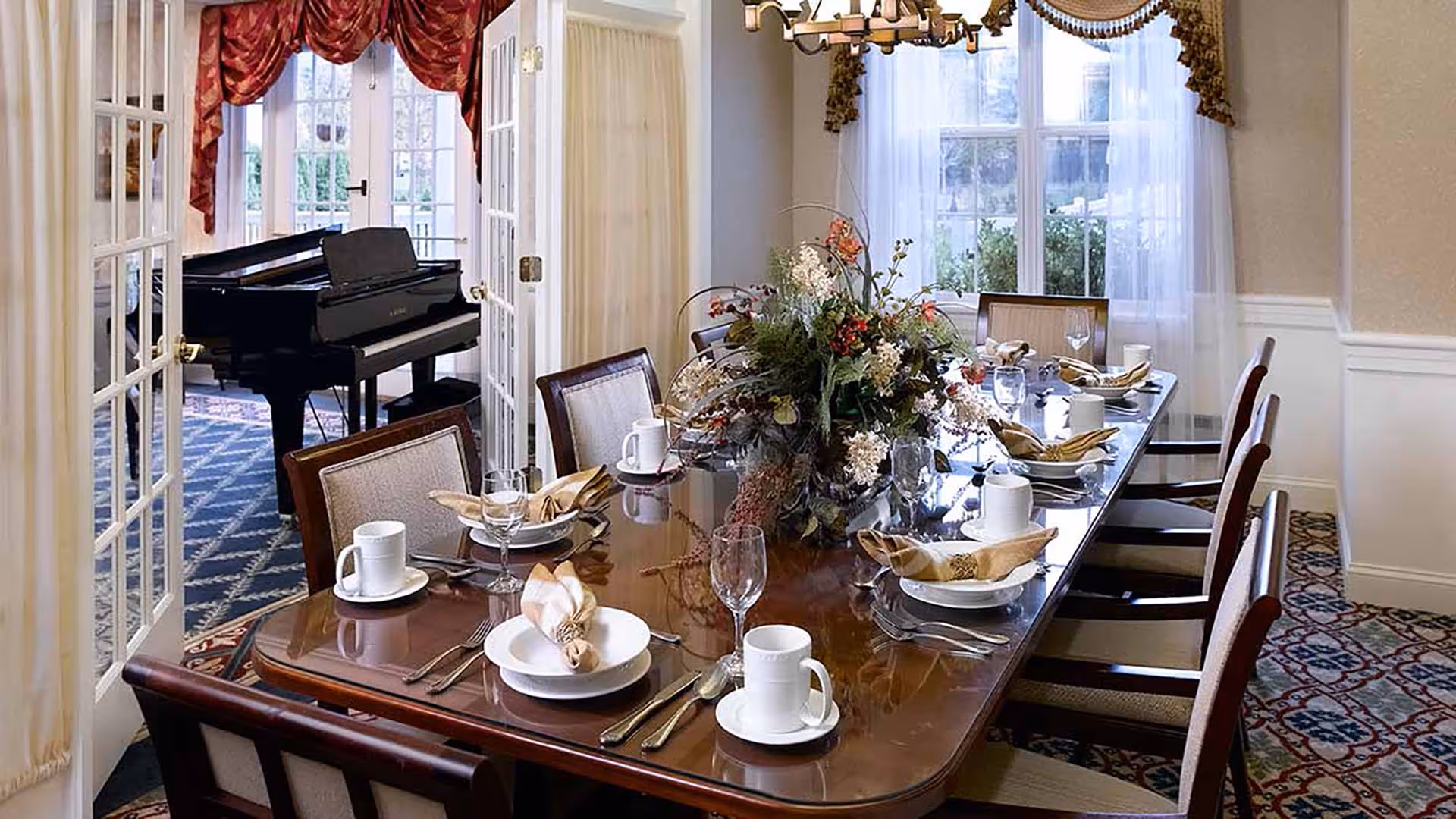 A formal dining room with a polished wooden table set for eight people, featuring white plates, beige napkins, glassware, and coffee mugs. A large floral centerpiece decorates the middle of the table. The room has large windows with sheer white curtains and ornate valances. In the adjacent room, a black grand piano is visible through glass-paneled double doors.