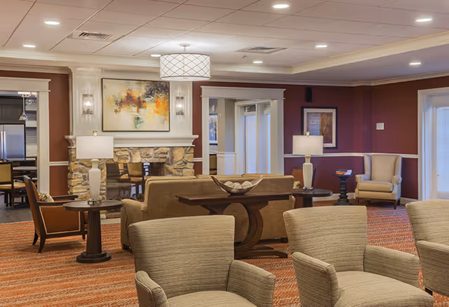 A cozy senior living facility common area with beige armchairs and sofas arranged around a stone fireplace. The room features warm lighting from table lamps and a ceiling light fixture, with artwork on the walls and a patterned carpet.