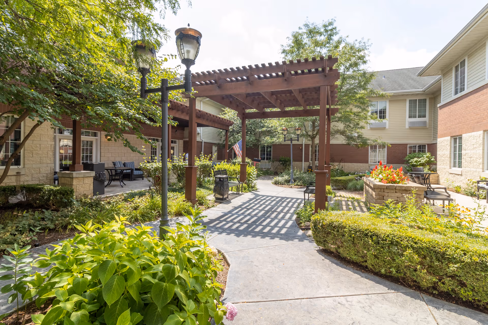 Outdoor courtyard area at StoryPoint Romeoville featuring a wooden pergola, lamp posts, benches, well-maintained shrubs, flowering plants, and a paved walkway surrounded by the building's exterior walls with windows and seating areas.