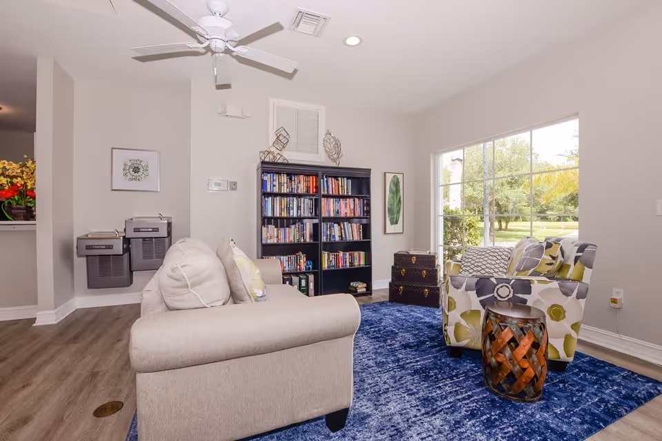 A cozy living room area with a beige sofa and a floral patterned armchair on a blue rug. There is a black bookshelf filled with books against the wall, a small decorative chest, and a large window showing a green outdoor view. The room has light-colored walls, wood flooring, a ceiling fan, and two water fountains mounted on the wall.