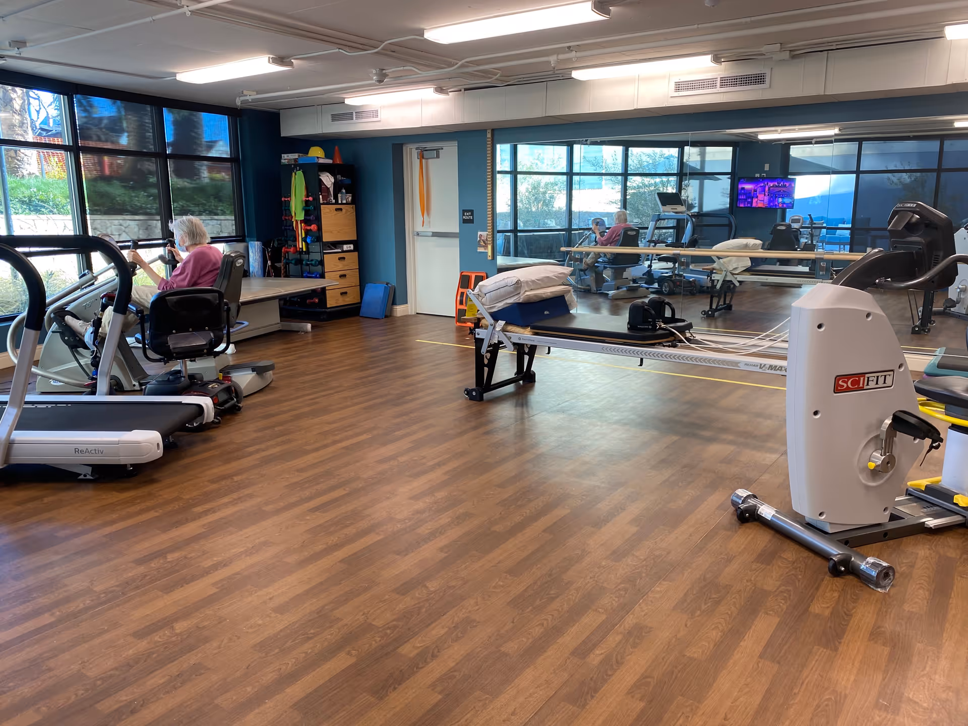 A fitness room with wooden flooring and large windows. An elderly person is using a seated exercise bike near the windows. The room contains various exercise equipment including a treadmill, a rowing machine, and other fitness apparatus. There is a large mirror on one wall reflecting the room and the person exercising. Shelves with exercise accessories and a door are visible in the background.