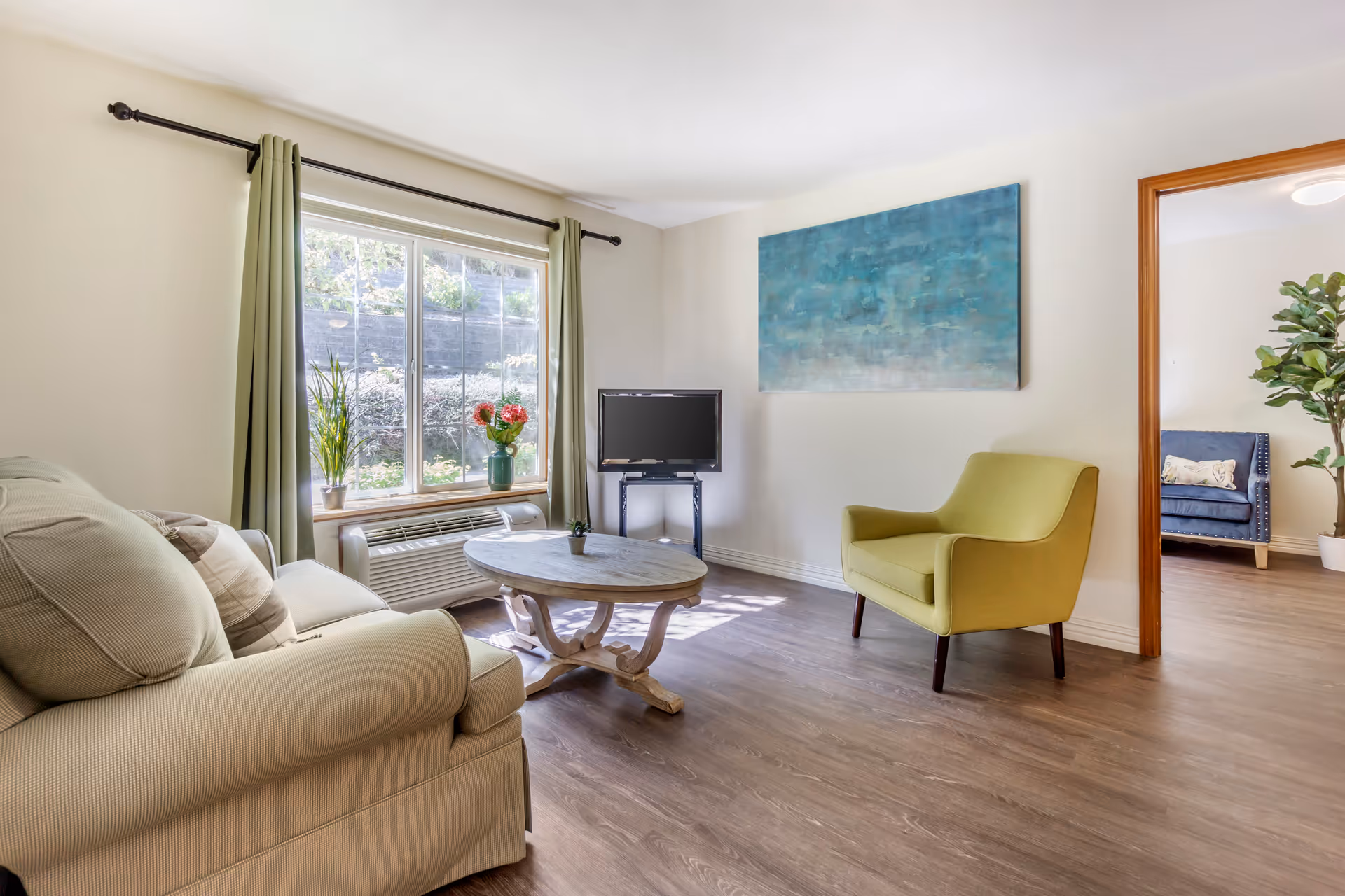 Bright furnished living room with a sofa, green armchair, round wooden coffee table, small TV, large window with green curtains, and abstract wall art.