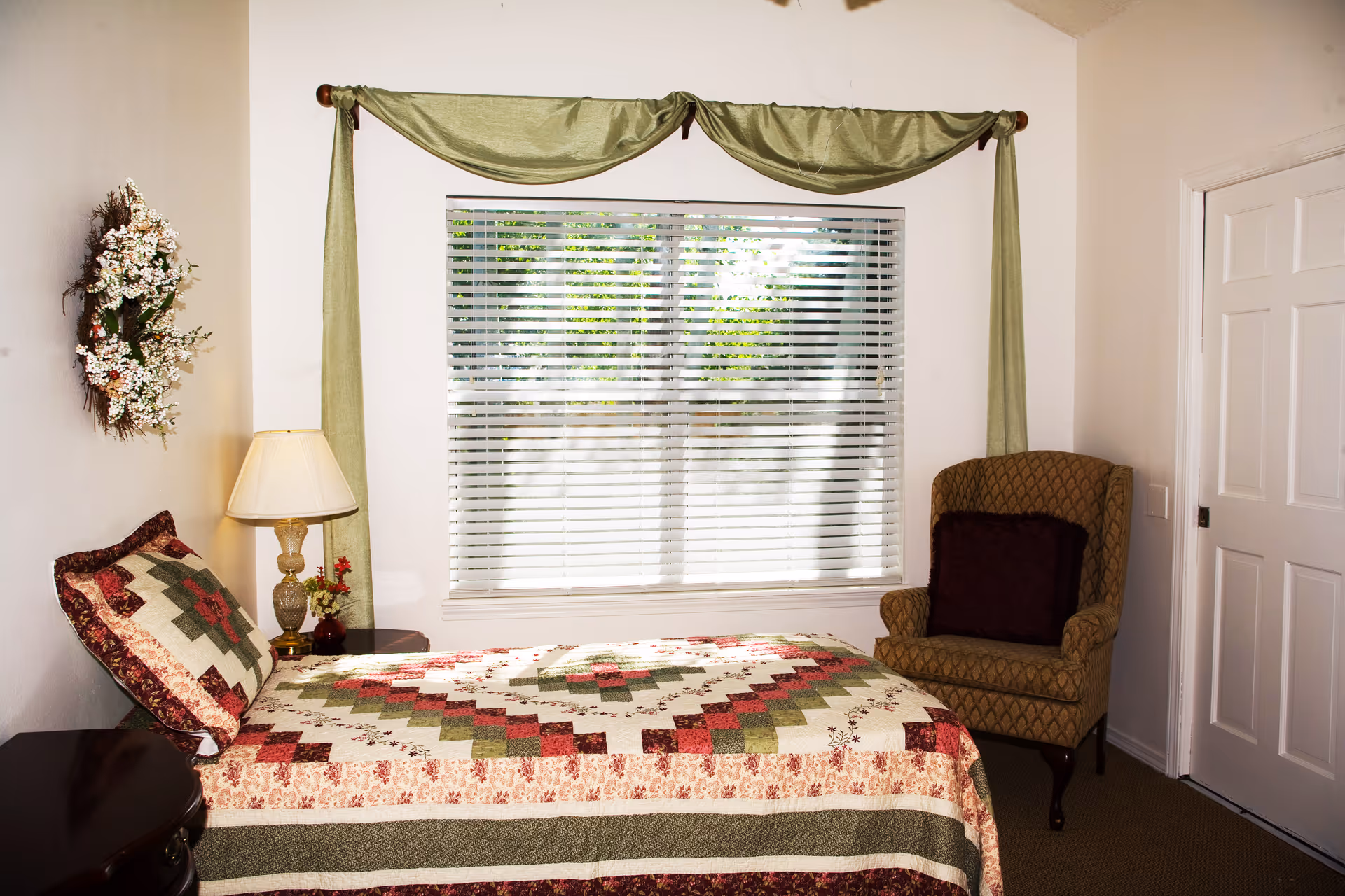 A cozy bedroom with a bed covered in a quilted bedspread featuring green, red, and cream patterns. Next to the bed is a wooden nightstand with a decorative lamp and a small vase with flowers. A cushioned armchair with a dark pillow is placed near a white door. The window has white blinds and green drapes, and a floral wreath hangs on the wall above the nightstand.