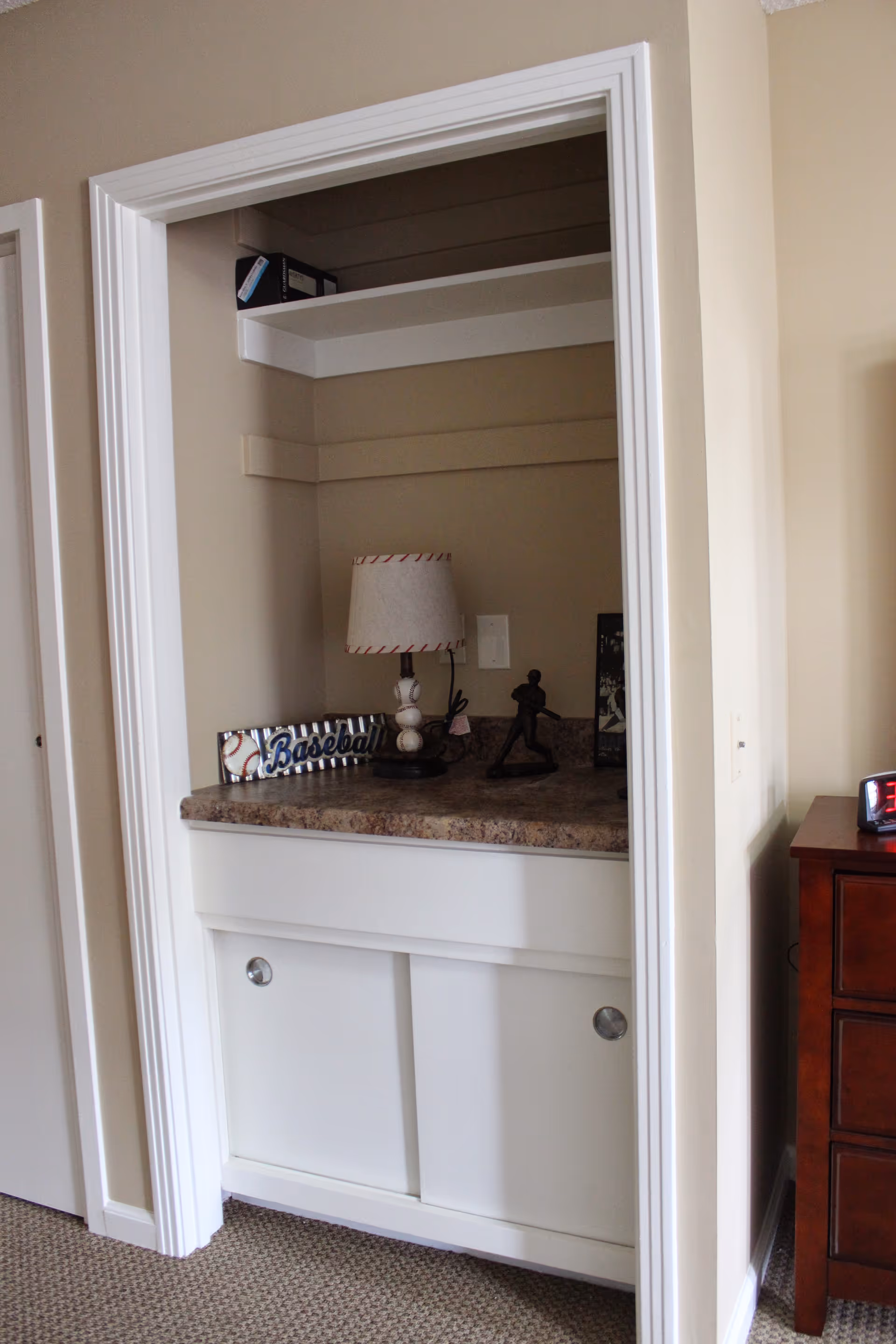 A small built-in nook with a countertop and sliding cabinet doors below. On the countertop are a table lamp, a decorative baseball sign, and a small baseball player figurine. The nook is framed by beige walls and white trim, with a carpeted floor visible in front.