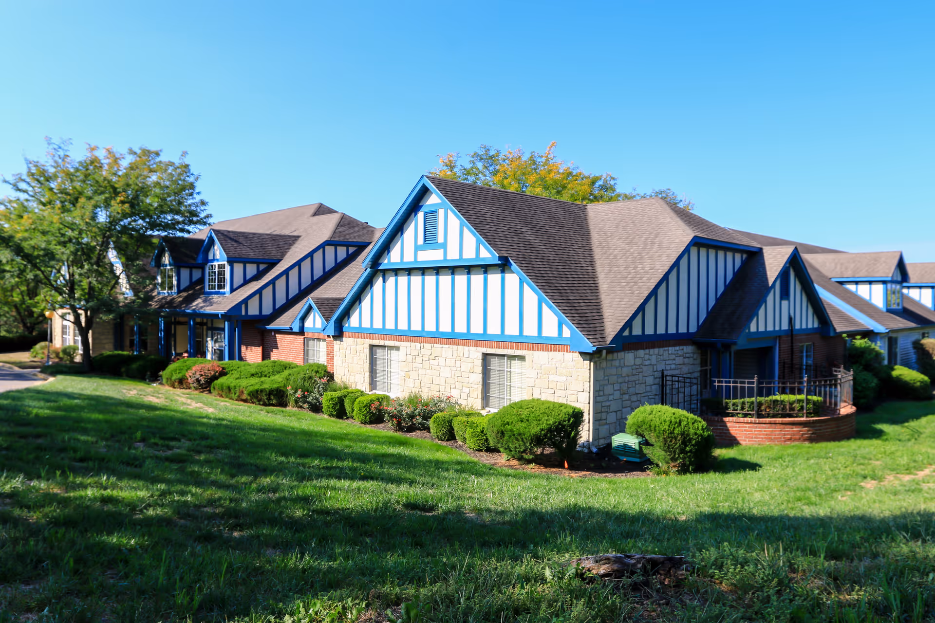 Exterior view of a residential building with Tudor-style architecture featuring blue and white trim, stone and brick walls, surrounded by green bushes and a grassy lawn under a clear blue sky.