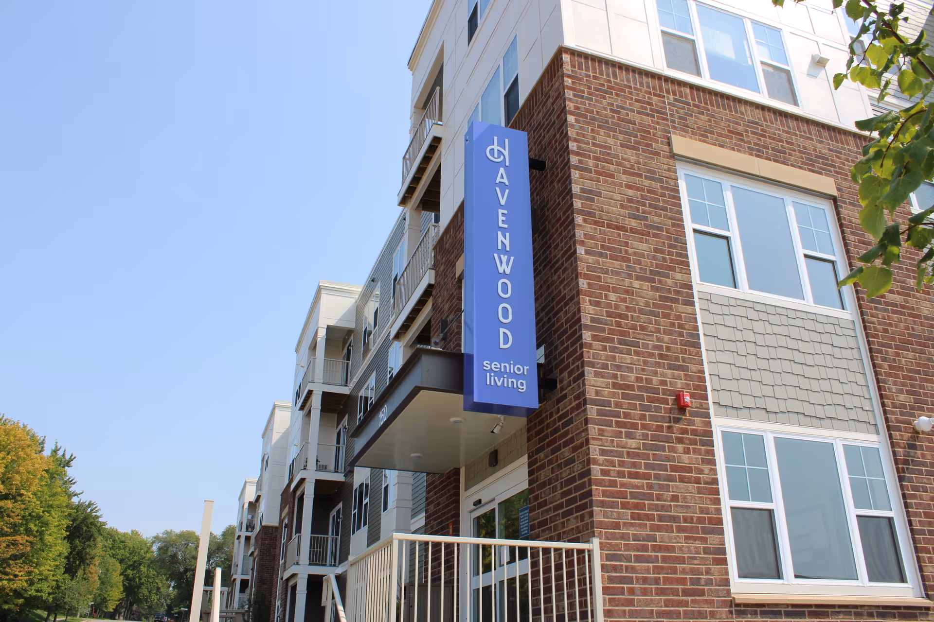 Exterior view of a multi-story senior living facility building with a brick and siding facade under a clear blue sky. A vertical blue sign on the building reads 'Havenwood senior living'. Trees and a street are visible in the background.