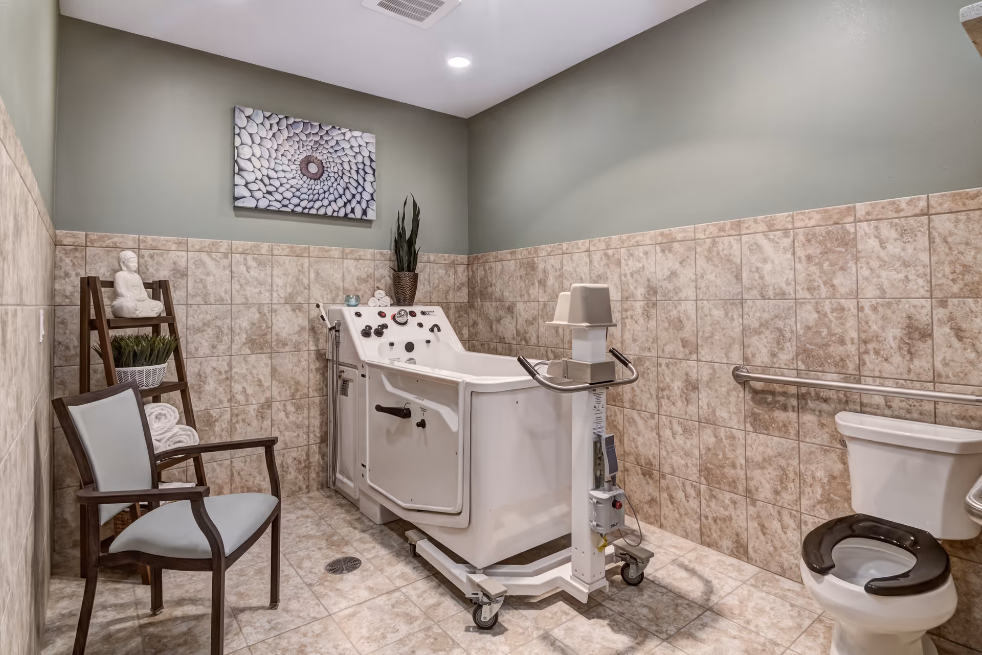 A tiled bathroom with a specialized assisted bathing tub on wheels, a toilet with a black seat, a wooden chair with light blue cushion, a wooden shelf with a white Buddha statue, rolled towels, and a potted plant. The walls are half-tiled with beige tiles and painted green above. A decorative artwork hangs on the wall above the tub.