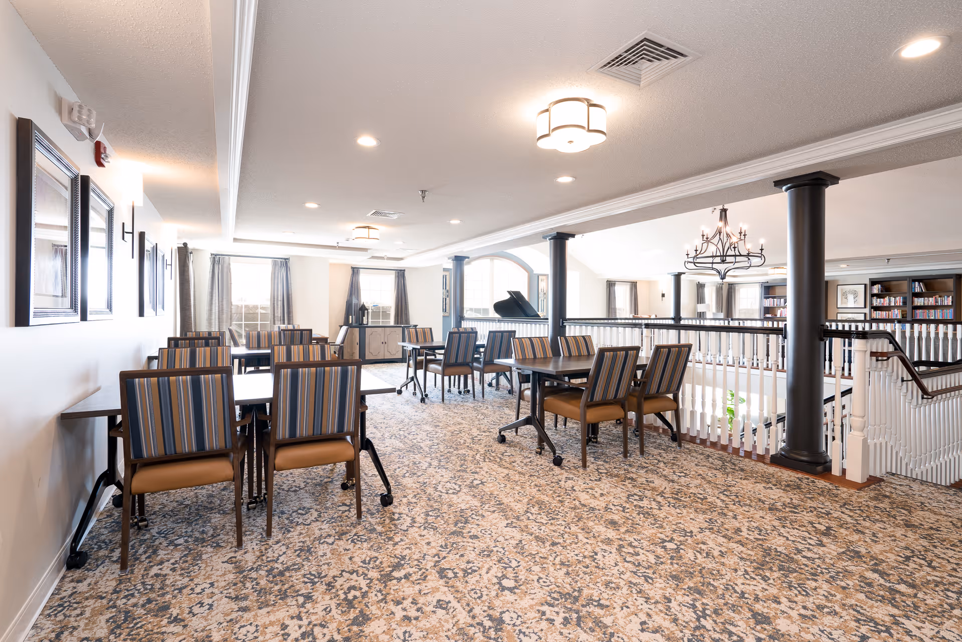 A bright and spacious common area with multiple tables and striped cushioned chairs arranged for seating. The room features large windows with curtains, a patterned carpet, white walls with framed artwork, and black columns supporting the ceiling. A grand piano and a bookshelf filled with books are visible in the background, along with a decorative chandelier hanging from the ceiling.