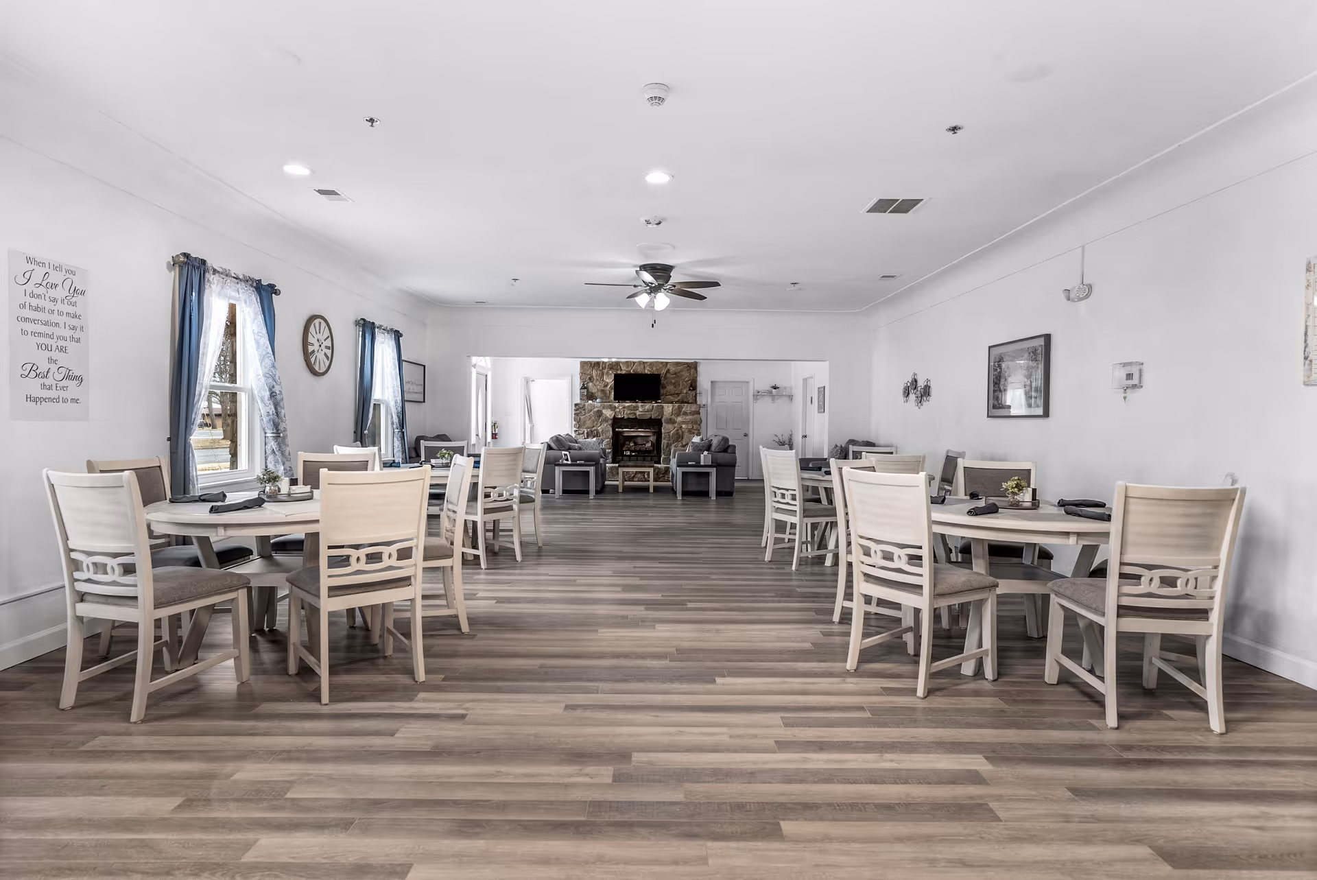 A spacious dining area in an assisted living facility with multiple round tables and chairs arranged neatly. The room has wooden flooring, white walls, and windows with blue curtains letting in natural light. At the far end, there is a stone fireplace with a TV mounted above it and a seating area with sofas. Ceiling fans and recessed lighting are visible on the ceiling.