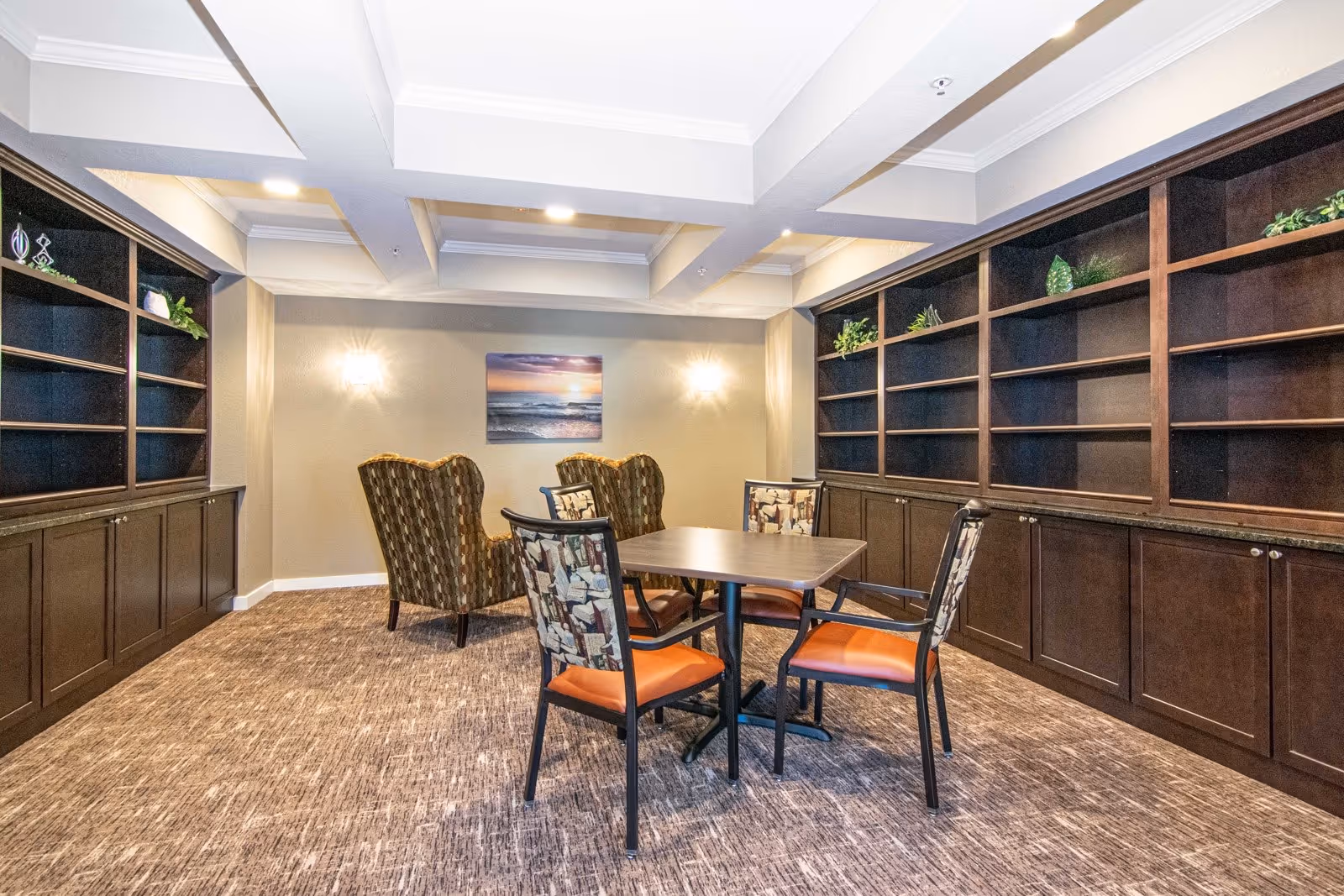 A cozy interior room with a square table surrounded by four chairs with patterned backs and orange seats. Behind the table are two cushioned armchairs with a patterned fabric. The walls are beige with two wall sconces and a framed picture of a sunset over the ocean. Dark wooden built-in shelves line both sides of the room, decorated with small plants and ornaments.