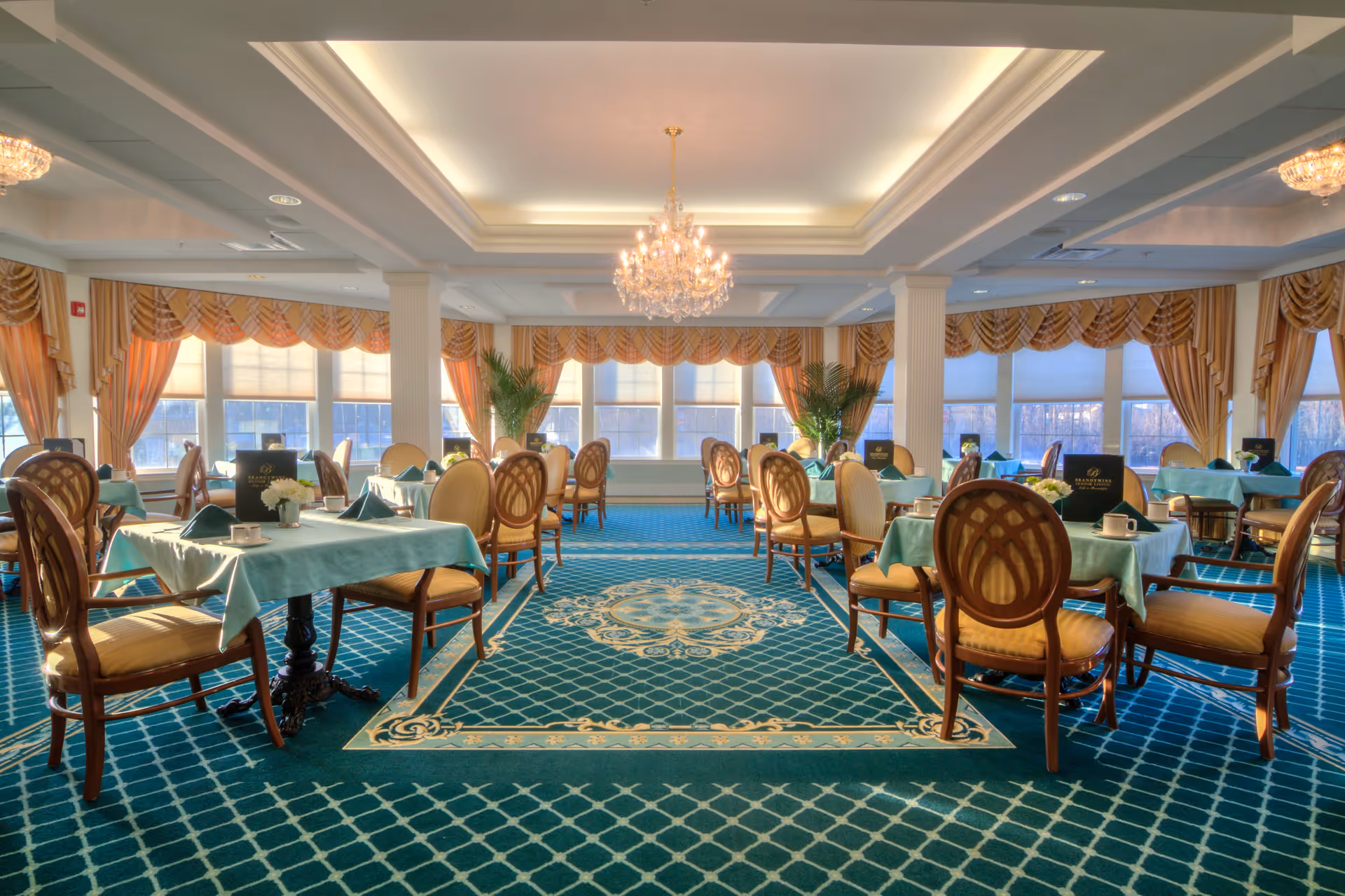 A spacious dining room with multiple tables covered in teal tablecloths, each set with napkins, cups, and small flower arrangements. The room features elegant wooden chairs with cushioned seats, large windows with beige and gold drapes, and a decorative blue carpet with a patterned design. A crystal chandelier hangs from the ceiling, adding a touch of sophistication to the room.