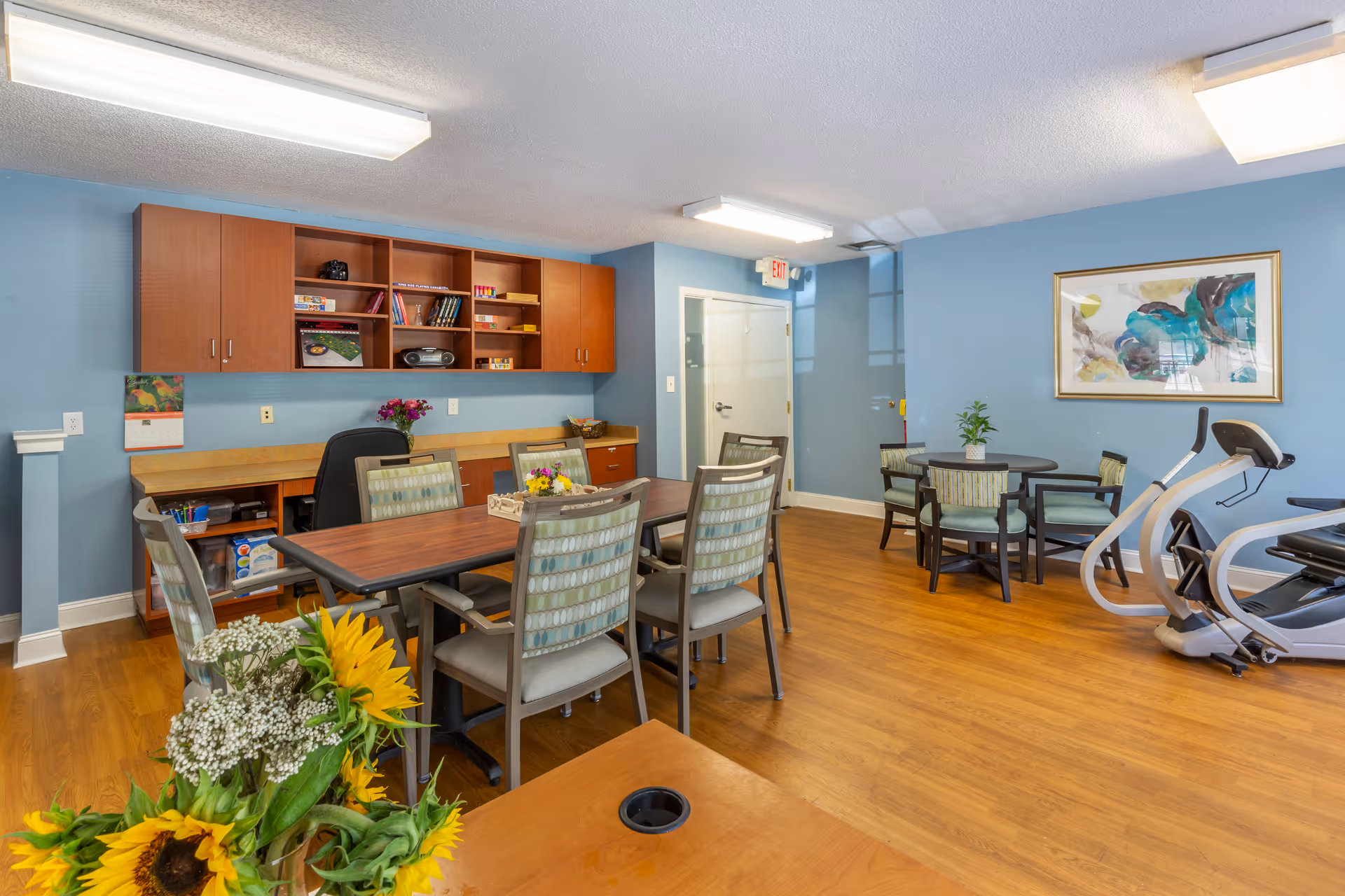 A bright room with light blue walls and wood flooring featuring two tables with chairs around them. One table has six chairs and the other has four chairs. There are flowers on the tables and a cabinet with shelves on the wall containing books, a radio, and games. An elliptical exercise machine is positioned near the smaller table. A framed abstract painting hangs on the wall above the smaller table. The room is lit by ceiling lights and has a door with an exit sign above it.