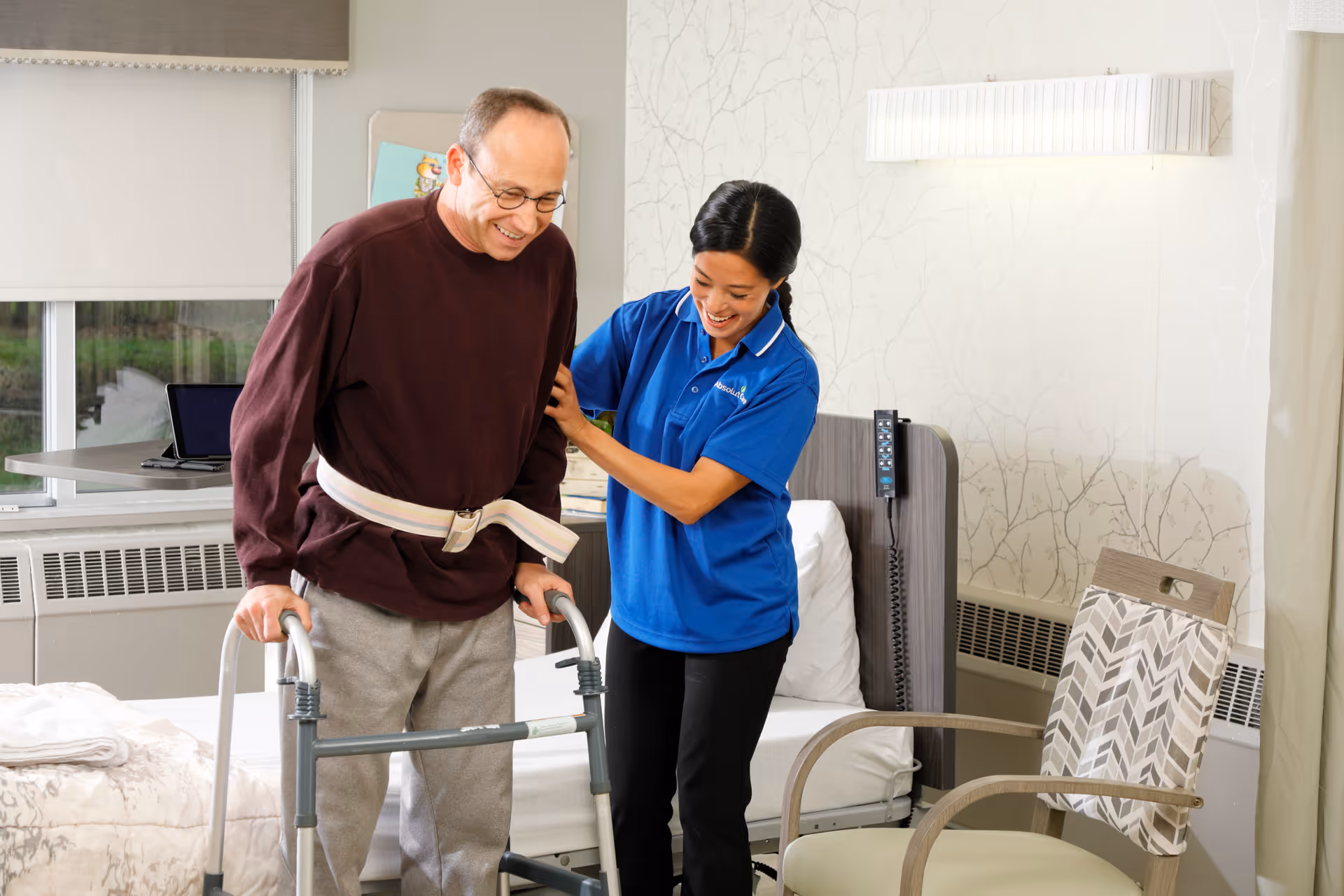 A male senior using a walker is assisted by a female caregiver wearing a blue Absolut Care polo shirt in a bedroom setting with a bed, chair, and window in the background.