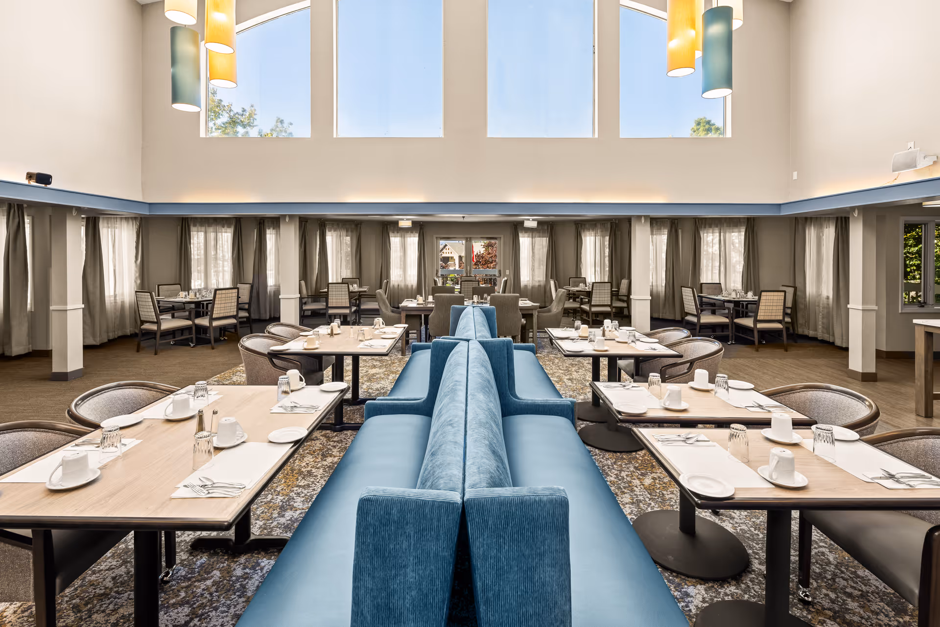 A spacious dining room with high ceilings and large windows letting in natural light. The room features multiple tables set with white cups, plates, and silverware. Blue cushioned bench seating runs down the center, flanked by individual chairs at each table. Modern pendant lights hang from the ceiling, and curtains cover the windows along the walls.