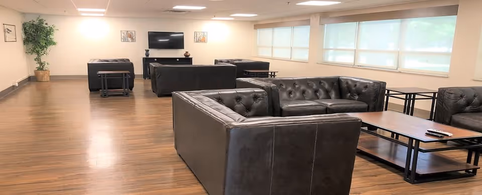 A spacious living room area with multiple black leather tufted sofas arranged around wooden coffee tables. The room has large windows with white blinds, a flat-screen TV mounted on the wall, wooden flooring, and a potted plant in the corner.