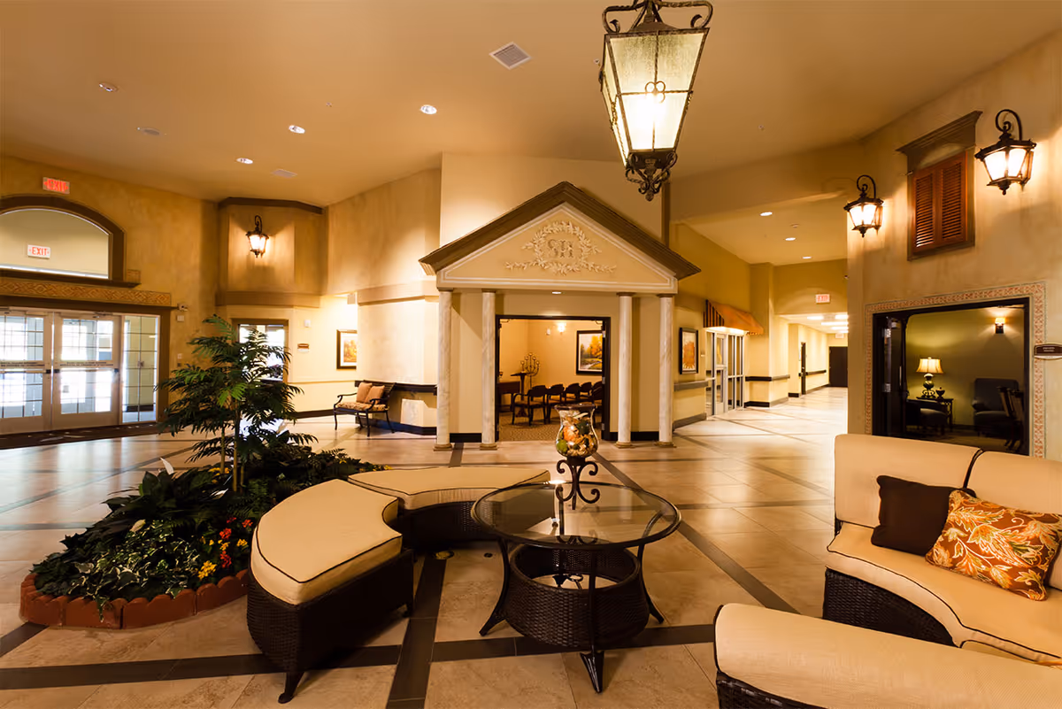 Spacious, warmly lit interior lobby with wicker seating, a glass coffee table, indoor plants, and decorative columns.
