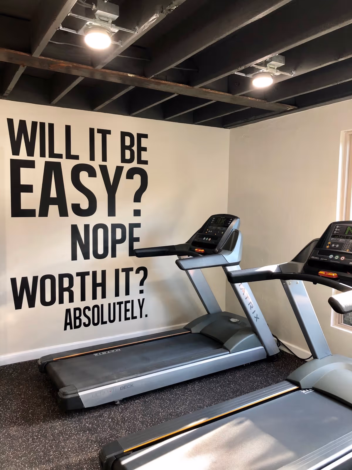 Indoor gym area with two treadmills positioned side by side. On the wall behind the treadmills, there is a motivational quote in large black letters that reads: 'WILL IT BE EASY? NOPE WORTH IT? ABSOLUTELY.' The ceiling has exposed beams and lighting fixtures.