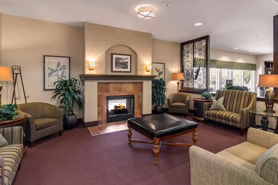 Cozy communal living room with a fireplace, upholstered chairs, a central ottoman, lamps, and plants.