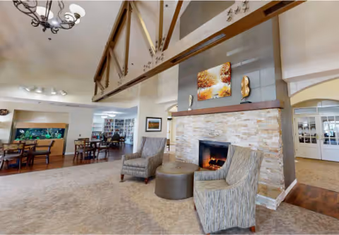 A cozy common area in a senior living facility featuring two patterned armchairs and a round ottoman in front of a stone fireplace with a fire burning. Above the fireplace is a colorful painting and decorative sculptures. The room has a high ceiling with exposed wooden beams and a chandelier. In the background, there are tables and chairs near a large aquarium and a bookshelf-filled room.