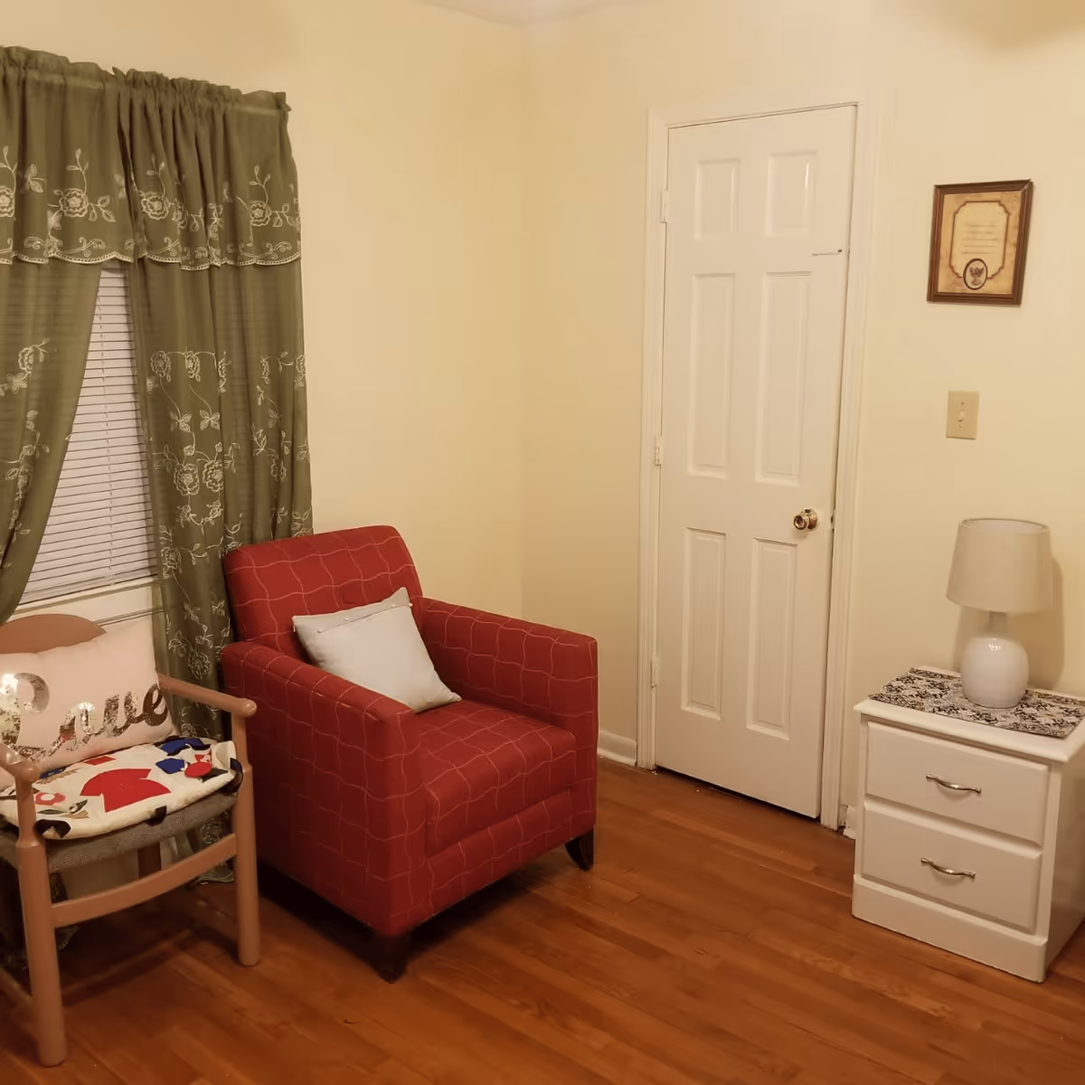 A cozy corner of a room with a red armchair featuring a light blue pillow, a wooden chair with a cushion and a pillow that says 'Love', green curtains with floral embroidery covering a window, a white door, and a white nightstand with two drawers and a lamp on top. The floor is wooden.