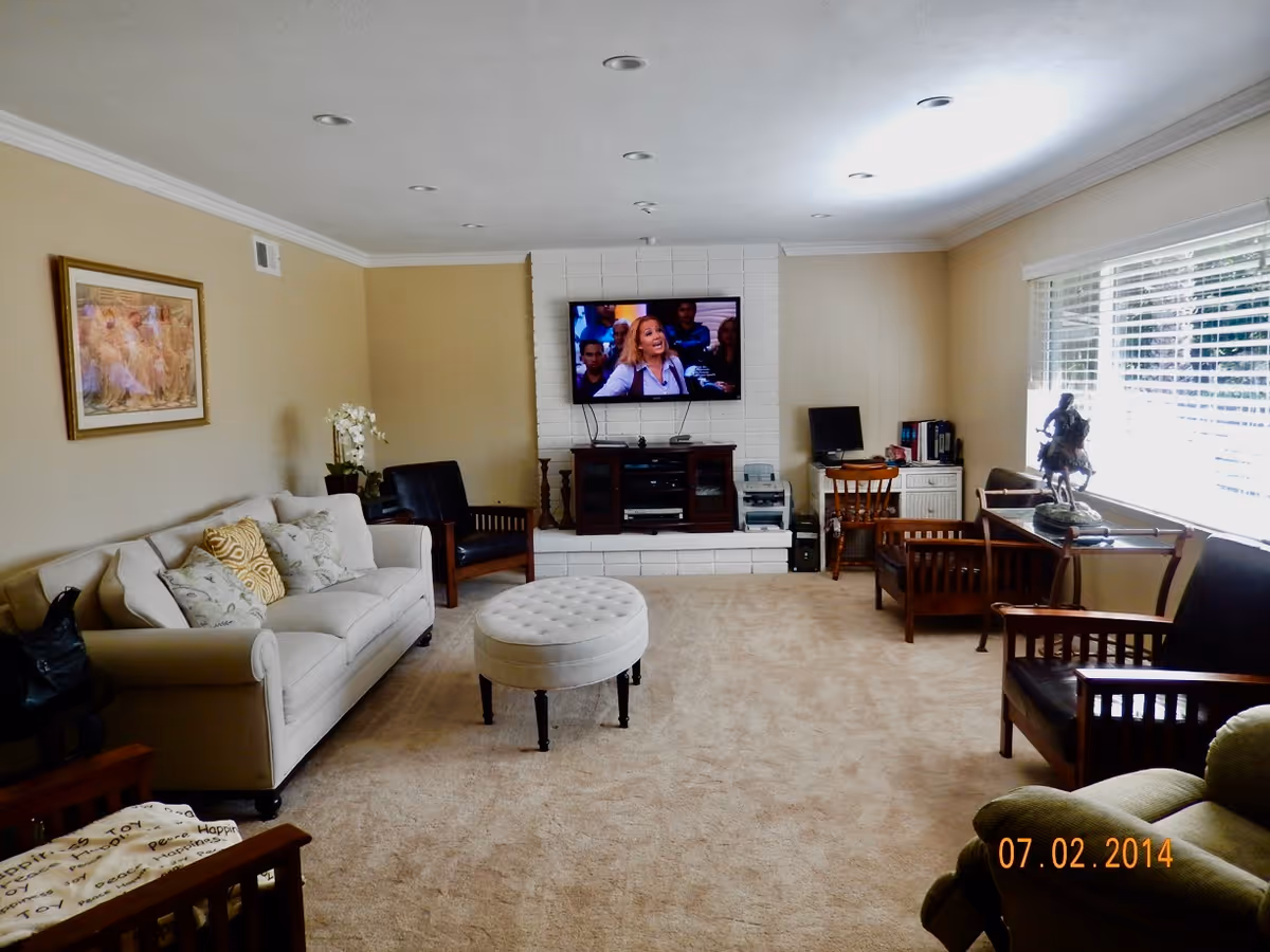 Bright living room with beige walls, a sofa and chairs facing a wall-mounted TV above a white brick hearth and a round ottoman in the center.
