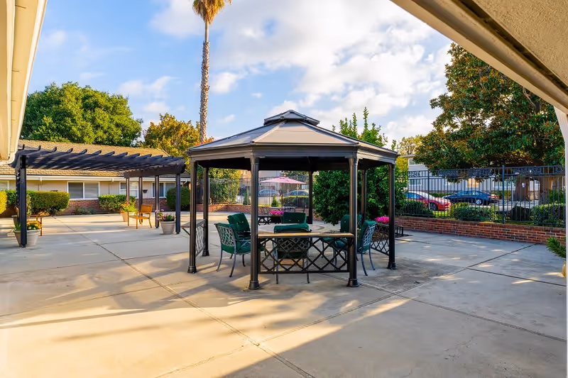 Outdoor patio area at Silver Oaks Memory Care featuring a gazebo with green cushioned chairs and a table underneath. Surrounding the patio are trees, bushes, and a pergola with benches. Cars and a fence are visible in the background under a partly cloudy sky.