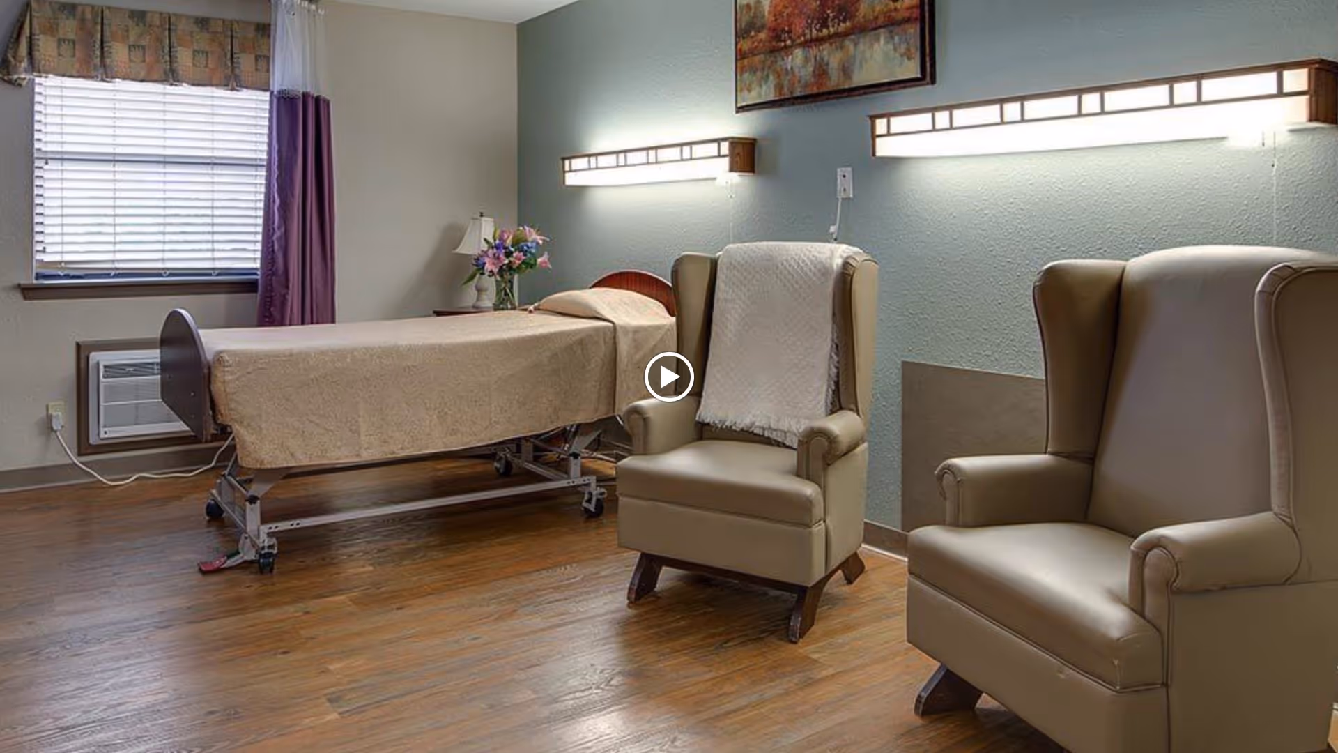 A nursing center room with a single hospital bed covered in a beige blanket, two beige armchairs with a white throw on one, a window with blinds and curtains, a small side table with a lamp and flowers, and two wall-mounted lights above the chairs.