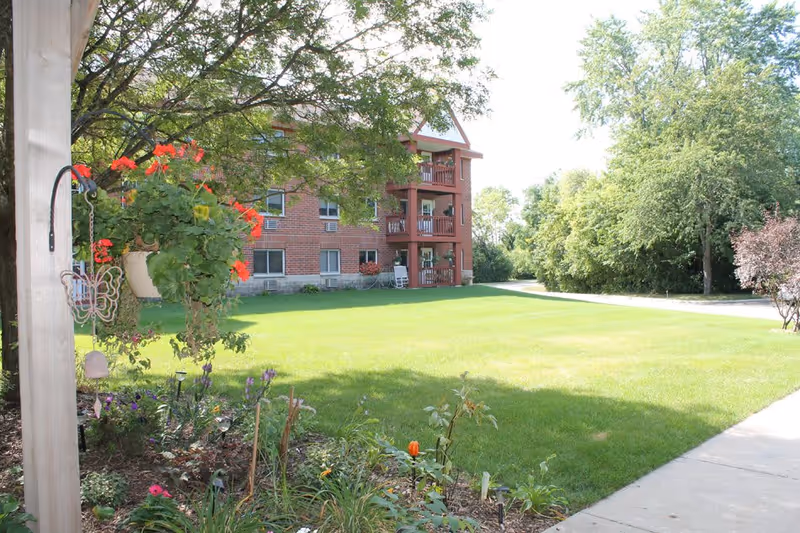 A sunny outdoor view of a green lawn with a flower garden in the foreground and a multi-story brick building with balconies in the background. Trees surround the area, and a sidewalk runs along the right side of the image.