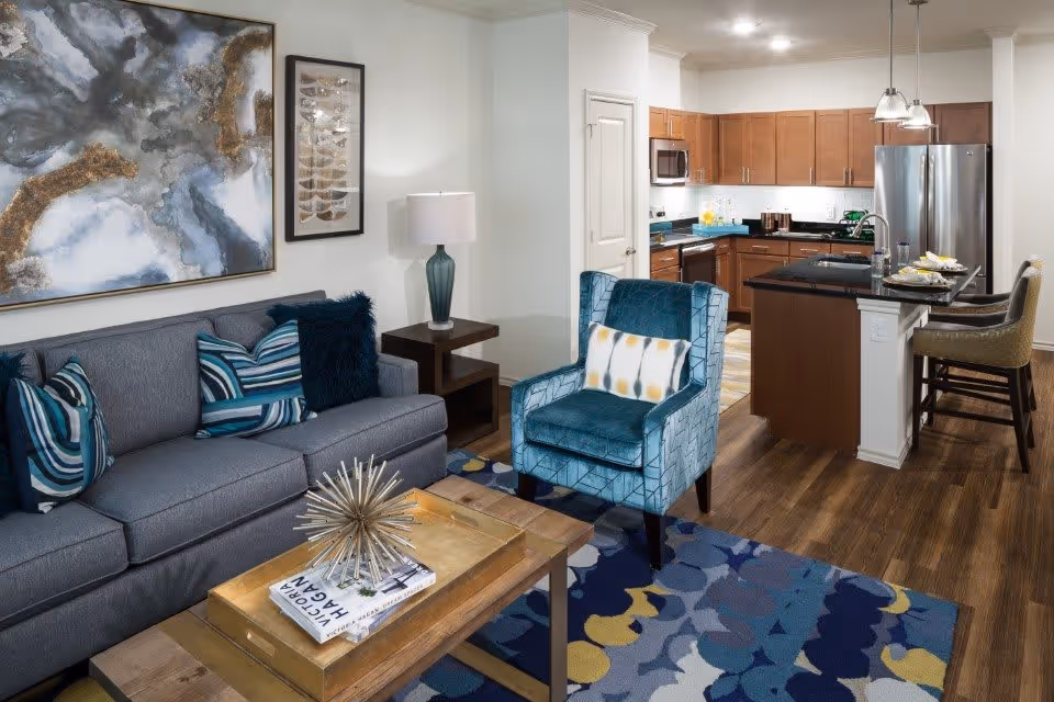 A modern living room and kitchen area in a senior living facility. The living room features a gray sofa with blue and striped pillows, a blue armchair with a patterned cushion, a wooden coffee table with decorative items, and a colorful abstract rug. The kitchen has wooden cabinets, stainless steel appliances, a black countertop island with two bar stools, and pendant lighting.