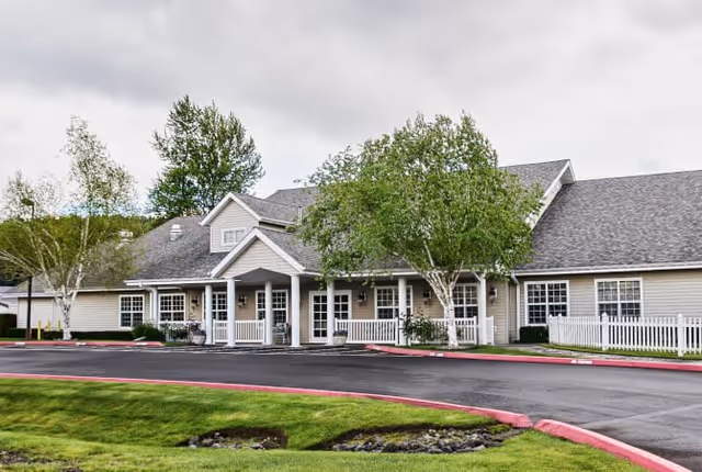 Exterior view of a single-story senior living facility building with beige siding, white trim, and a gray shingled roof. The entrance features a covered porch with white railings and columns. There are several windows along the building, green trees, and a well-maintained lawn with a curved driveway in front.