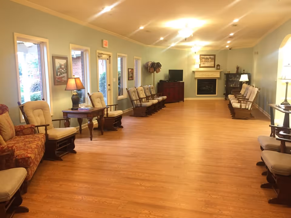 Spacious communal living room with wood floors, rows of cushioned rocking chairs, side tables and lamps, and a fireplace at the far end.