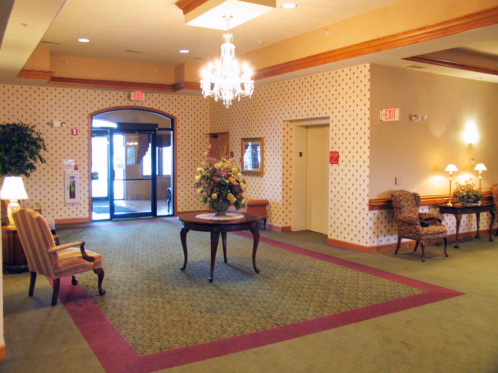 A spacious and well-lit lobby area with patterned wallpaper and carpet. There is a round wooden table with a floral arrangement in the center of the room. Several upholstered chairs are placed around the perimeter, along with side tables holding lamps and decorative plants. An elevator is visible on the right side, and glass double doors leading outside are at the far end. A chandelier hangs from the ceiling, adding an elegant touch.