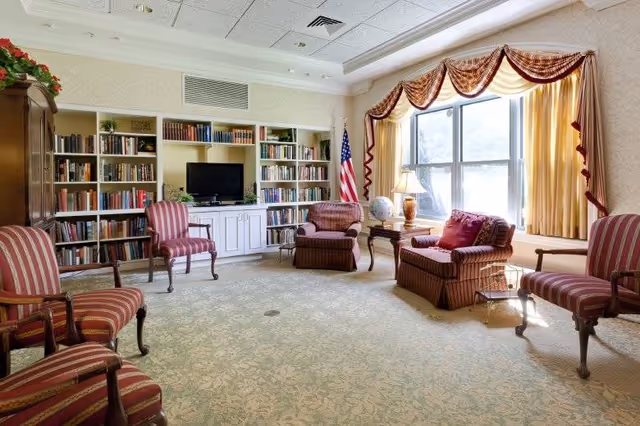 A cozy living room with several upholstered chairs arranged around the perimeter. There is a large window with yellow curtains and valances letting in natural light. A bookshelf filled with books and a TV is built into the wall. An American flag stands in the corner next to a side table with a lamp and a globe.