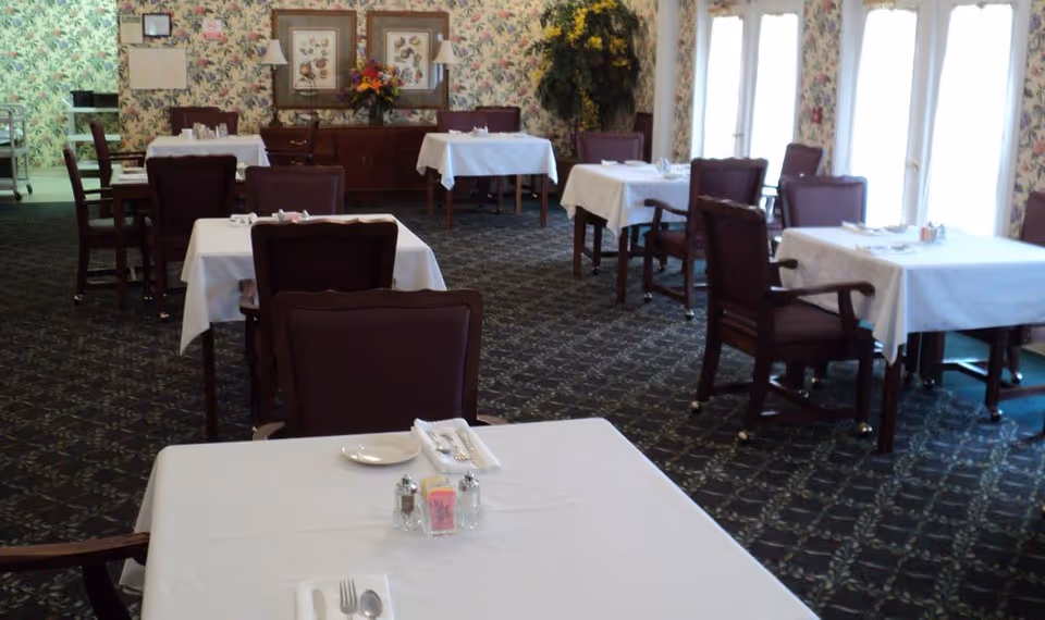 A dining room with several tables covered in white tablecloths, each set with plates, utensils, and condiments. The room has patterned carpet and floral wallpaper, with framed artwork and a flower arrangement on a sideboard against the wall. Large windows allow natural light to enter the space.