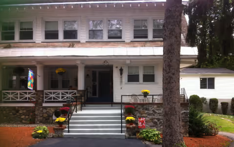 White two-story nursing facility with front porch, steps, and potted flowers.