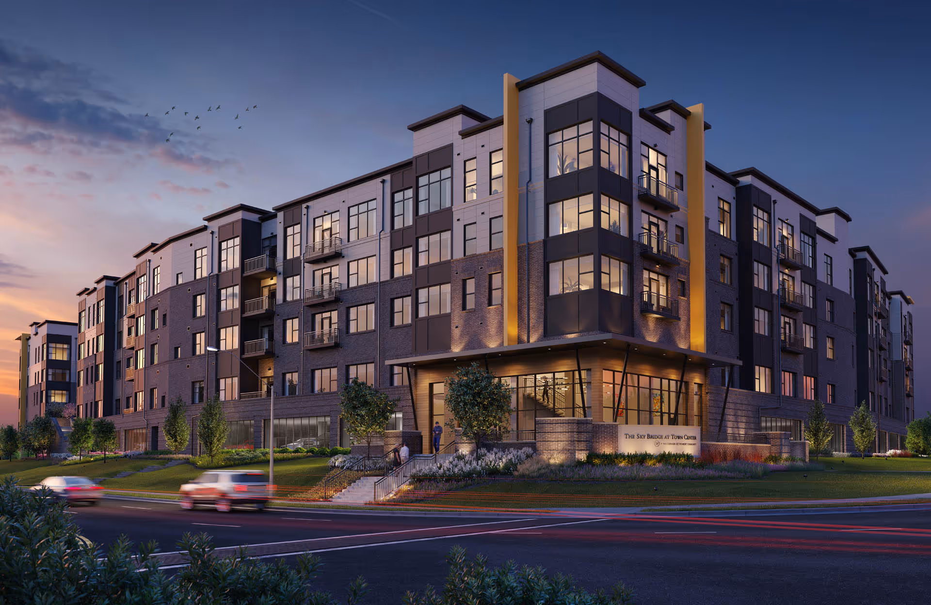 Exterior view of a modern multi-story senior living facility named The Sky Bridge at Town Center during dusk, with lights glowing from the windows and cars passing by on the street in front.